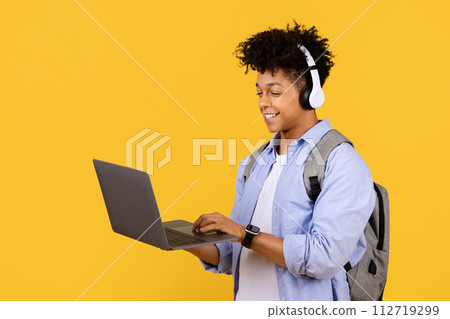 Happy student guy with headphones using laptop on yellow background Happy student guy with headphones using laptop on yellow background 112719299