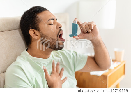 Handsome black man sitting alone at home, using asthma pump 112719319