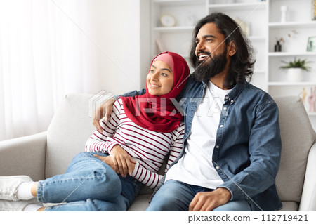 Happy muslim couple in casual wear sitting closely on couch and smiling 112719421