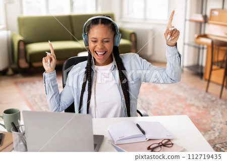 Joyful black teen student with headphones dancing at her desk Joyful black teen student with headphones dancing at her desk 112719435