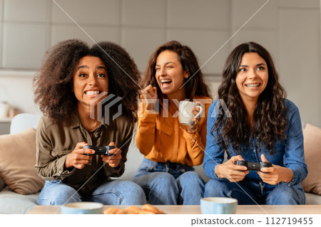 Multicultural female friends playing video games in their living room Multicultural female friends playing video games in their living room 112719455