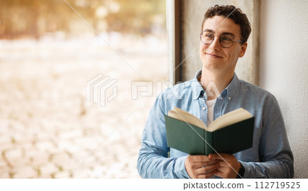 A contented young man with curly hair and round glasses is engrossed in reading a book 112719525