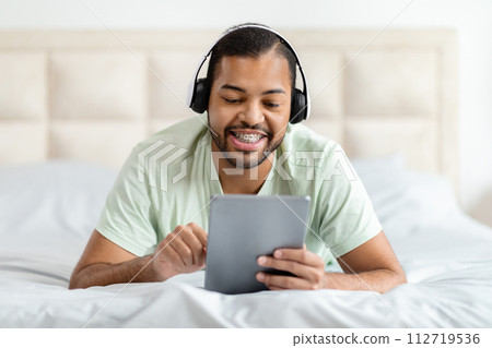 Happy black man using digital tablet and headphones in bed Happy black man using digital tablet and headphones in bed 112719536