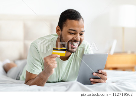 Black guy lying on bed, using digital tablet, bank card 112719562