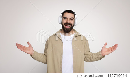Joyful young man with a beard wearing headphones and a beige overshirt over a white tee 112719614