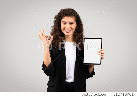 Cheerful businesswoman holding tablet with blank screen, making okay gesture 112719761