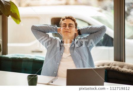 Relaxed and content young man with curly hair and glasses leaning back with hands behind his head 112719812