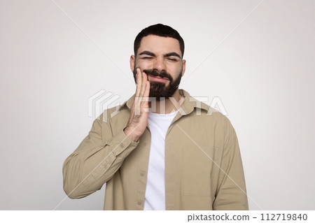 Man with a beard winces in discomfort, touching his cheek with a hand 112719840