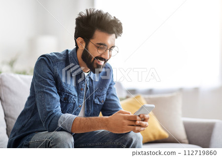 Cheerful bearded indian man looking at his smartphone with smile at home Cheerful bearded indian man looking at his smartphone with smile at home 112719860