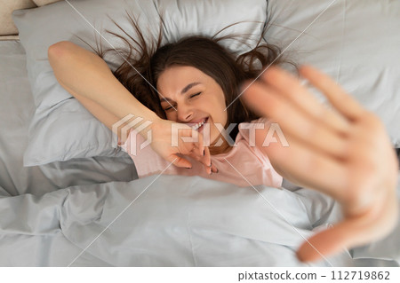 Woman waking up, stretching hand to camera and smiling, lying on bed in bedroom Woman waking up, stretching hand to camera and smiling, lying on bed in bedroom 112719862