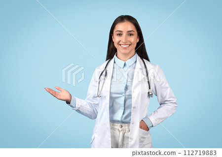 Smiling woman doctor presenting something with hand on blue backdrop Smiling woman doctor presenting something with hand on blue backdrop 112719883