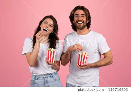 Joyful woman and man in white t-shirts laughing and eating popcorn from red and white striped boxes Joyful woman and man in white t-shirts laughing and eating popcorn from red and white striped boxes 112719893