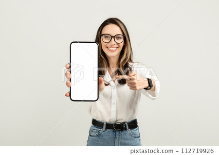 Engaging young woman showcasing a smartphone with a pointing gesture, wearing glasses 112719926