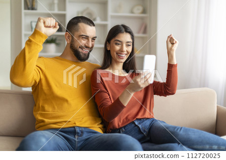 Happy Young Couple Celebrating Success While Looking At Smartphone Screen At Home 112720025