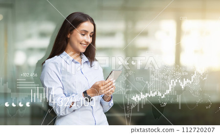 Young businesswoman in a light blue shirt uses her smartphone, with digital financial graphics 112720207