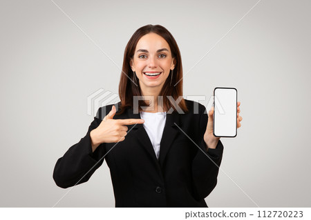 Enthusiastic businesswoman in a sleek black suit happily presenting a smartphone with a blank screen Enthusiastic businesswoman in a sleek black suit happily presenting a smartphone with a blank screen 112720223