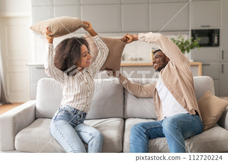 Carefree African American couple flirting on couch fighting with pillows Carefree African American couple flirting on couch fighting with pillows 112720224