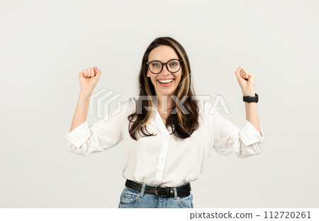 Joyful woman with glasses raising fists in excitement, wearing a white shirt and blue jeans Joyful woman with glasses raising fists in excitement, wearing a white shirt and blue jeans 112720261