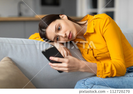 Worried young woman lying on couch looking at phone 112720270