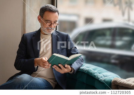 Thoughtful senior man with glasses engrossed in reading a book, comfortably seated 112720305