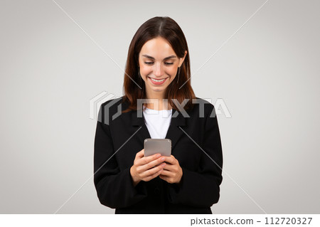 Content businesswoman in a classic black suit, engaged with her smartphone 112720327