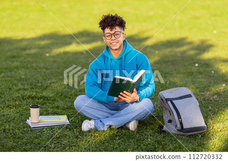 Cheerful black student guy in eyeglasses reading book at park 112720332