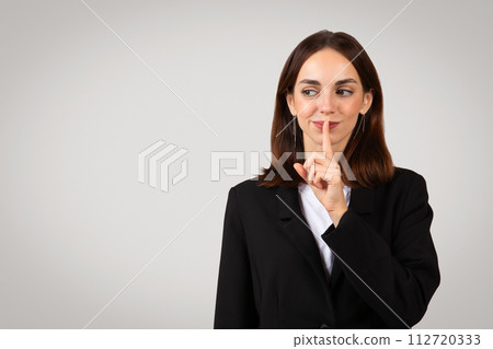 Playful businesswoman with a mischievous grin placing her finger over lips in a 'shush' gesture 112720333