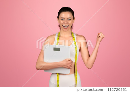Exultant young woman celebrating her weight loss success, holding a scale and wearing a measuring tape Exultant young woman celebrating her weight loss success, holding a scale and wearing a measuring tape 112720361