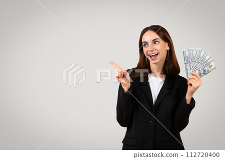 Excited businesswoman in a black suit holding a fan of dollar bills and pointing to the side Excited businesswoman in a black suit holding a fan of dollar bills and pointing to the side 112720400
