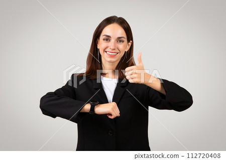 Cheerful businesswoman in a suit giving a thumbs up while checking the time on her smartwatch 112720408