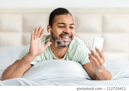 Cheerful young black guy lying on bed, have video call Cheerful young black guy lying on bed, have video call 112720411
