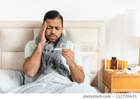 Unhealthy african american guy sitting in bed, checking fever 112720413
