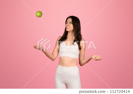 Playful young woman in white fitness attire joyfully tossing a green apple into the air 112720414