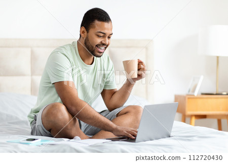 Relaxed black guy using laptop, sitting on bed Relaxed black guy using laptop, sitting on bed 112720430