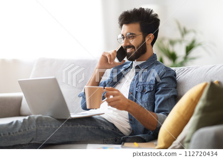 Indian man making phone call and holding coffee while working on laptop 112720434