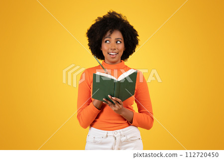 Inquisitive young woman with afro hair, in orange turtleneck, pensively holding a green book Inquisitive young woman with afro hair, in orange turtleneck, pensively holding a green book 112720450