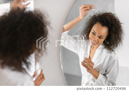 black woman doing haircare applying serum to hair in bathroom 112720486