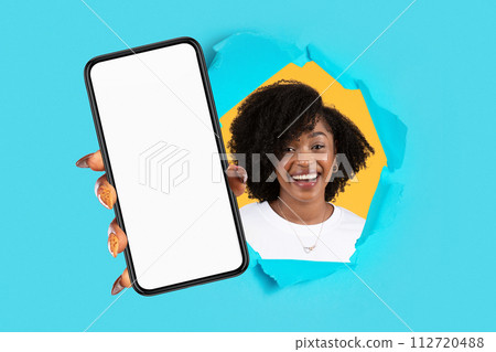 Smiling millennial african american lady showing smartphone with empty screen through hole in paper 112720488