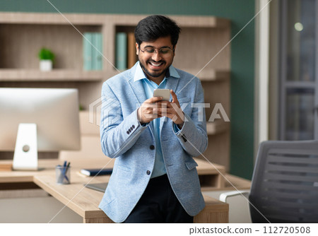 Indian CEO businessman browsing on his smartphone in modern office Indian CEO businessman browsing on his smartphone in modern office 112720508