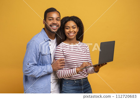 Young african american couple using laptop together while standing against yellow background 112720683