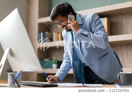 Indian businessman in conversation over phone taking notes in office 112720703