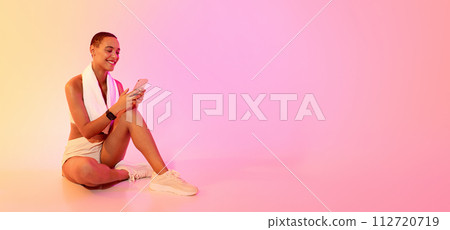 Joyful woman sitting cross-legged on the floor, looking at her smartphone with a smile 112720719