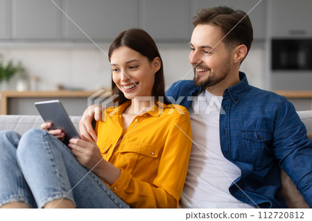 Couple smiling at tablet screen, resting on couch at home Couple smiling at tablet screen, resting on couch at home 112720812