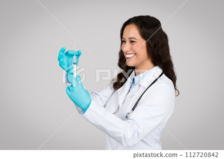 Cheerful young female doctor in white lab coat and gloves carefully preparing syringe for vaccination 112720842