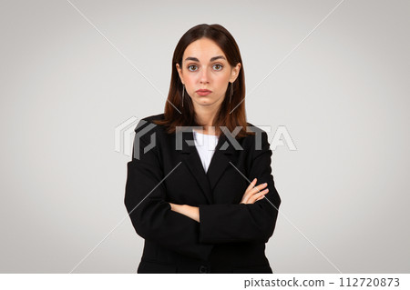Confident young businesswoman with long brown hair and a black suit stands with crossed arms 112720873