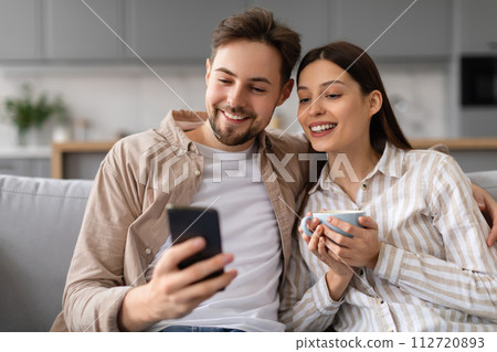 Spouses smiling at phone with coffee on couch 112720893