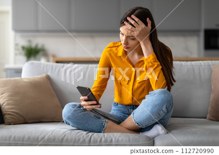 Worried young woman with phone, frowning on sofa 112720990