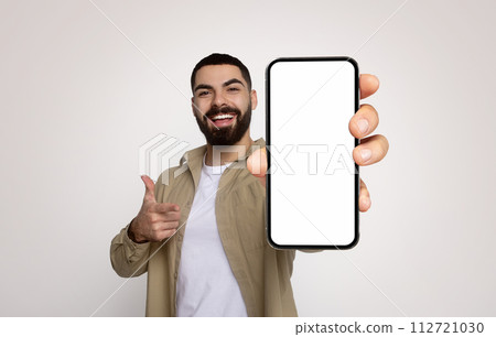 Enthusiastic young man with a beard, in a khaki shirt, cheerfully holds a smartphone with a blank screen 112721030