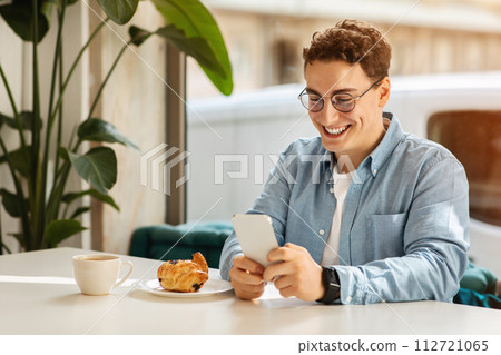 Happy young man with curly hair and glasses using his smartphone at a cafe table with a fresh croissant Happy young man with curly hair and glasses using his smartphone at a cafe table with a fresh croissant 112721065