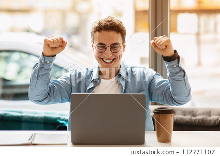 Joyful young man with curly hair wearing glasses and a blue shirt celebrates success Joyful young man with curly hair wearing glasses and a blue shirt celebrates success 112721107
